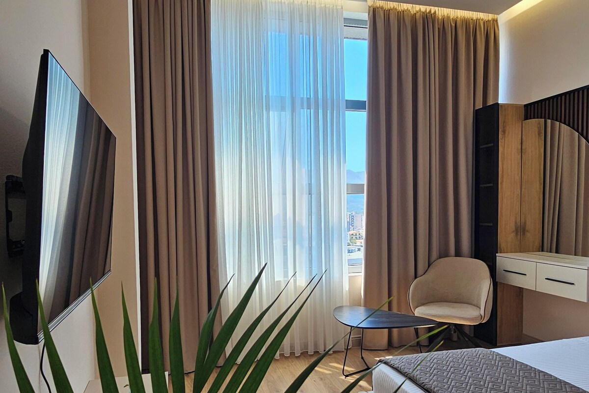 A modern hotel room with a bed, a desk, a chair, a TV, and a large window with curtains.