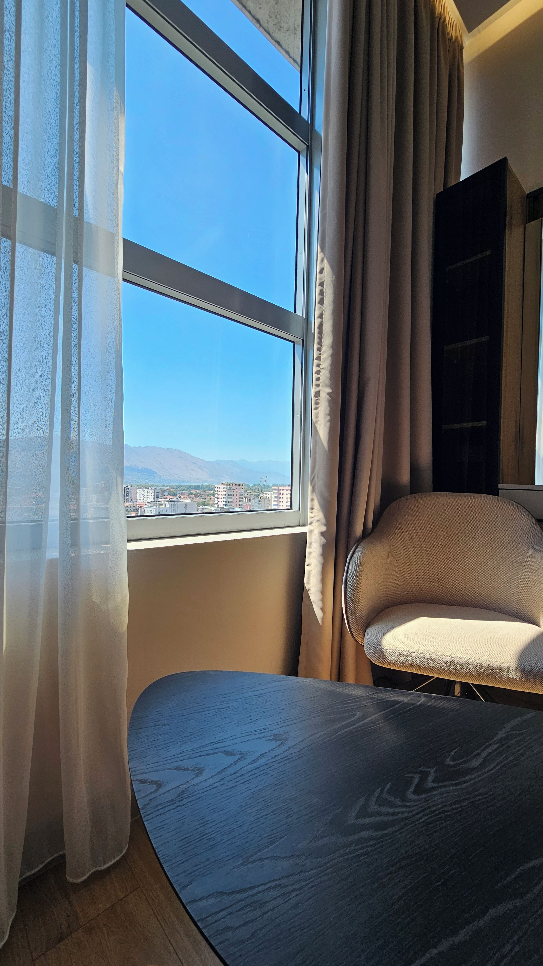 A room with a bed, a chair, a window with curtains, and a view of buildings and mountains.