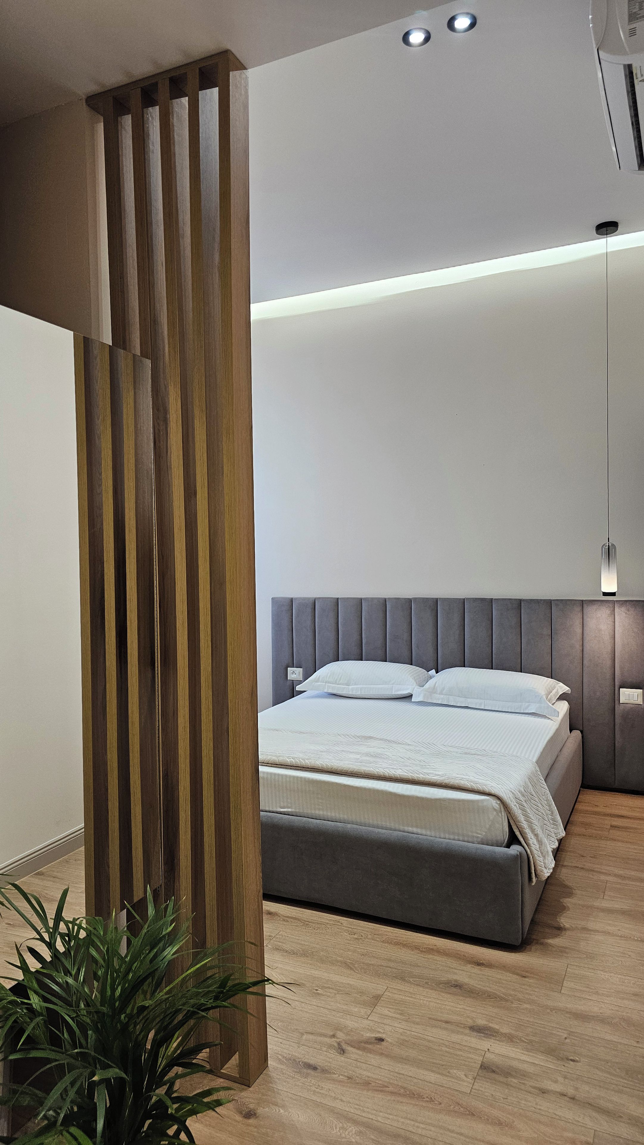 Een moderne slaapkamer met een grijs gestoffeerd hoofdeinde, een bed met wit beddengoed, een houten lattenwand en een hangende hanglamp.