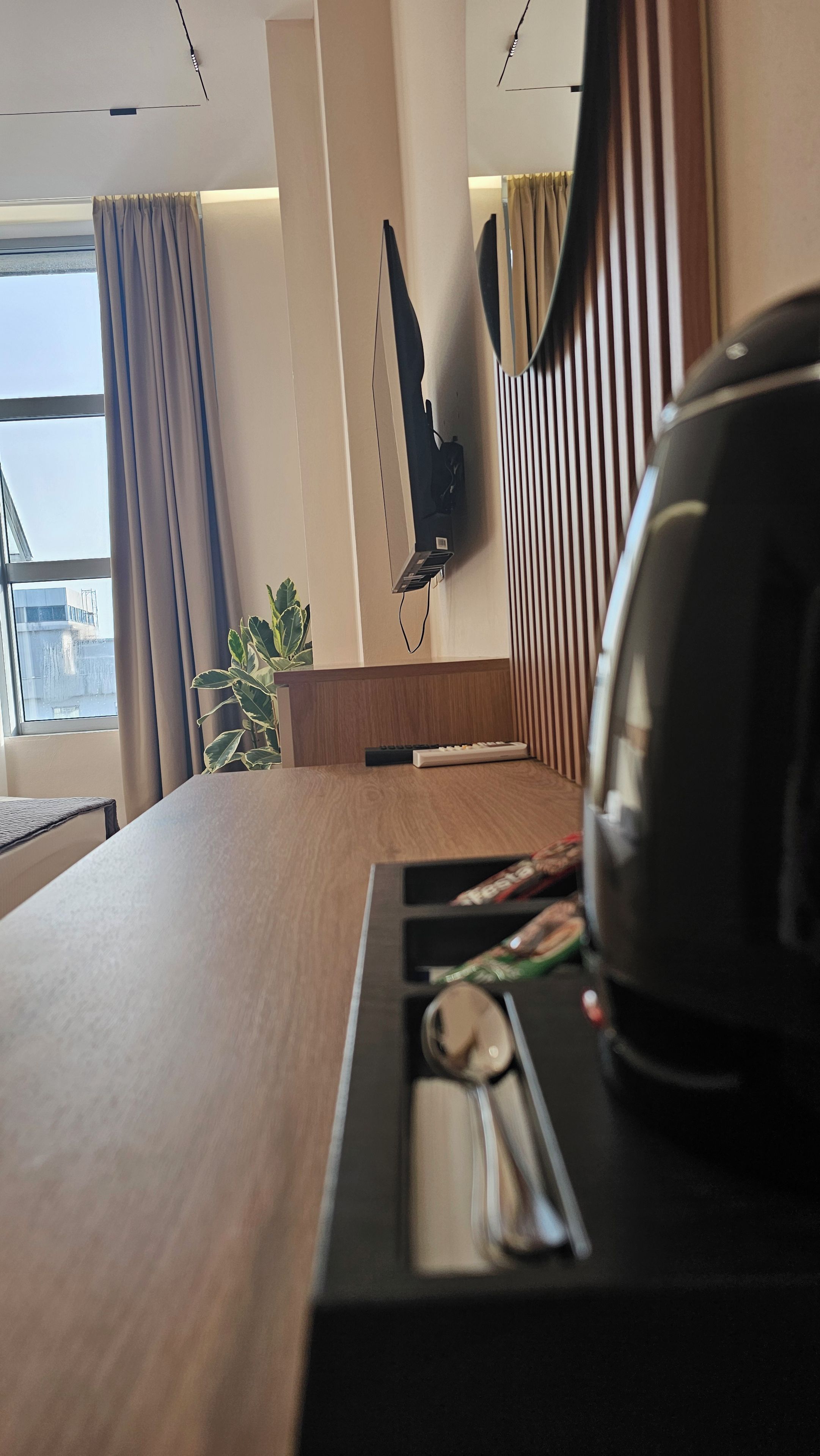 A modern hotel room with a wooden desk, a flat-screen TV mounted on the wall, a potted plant, and a view of the cityscape through the window.