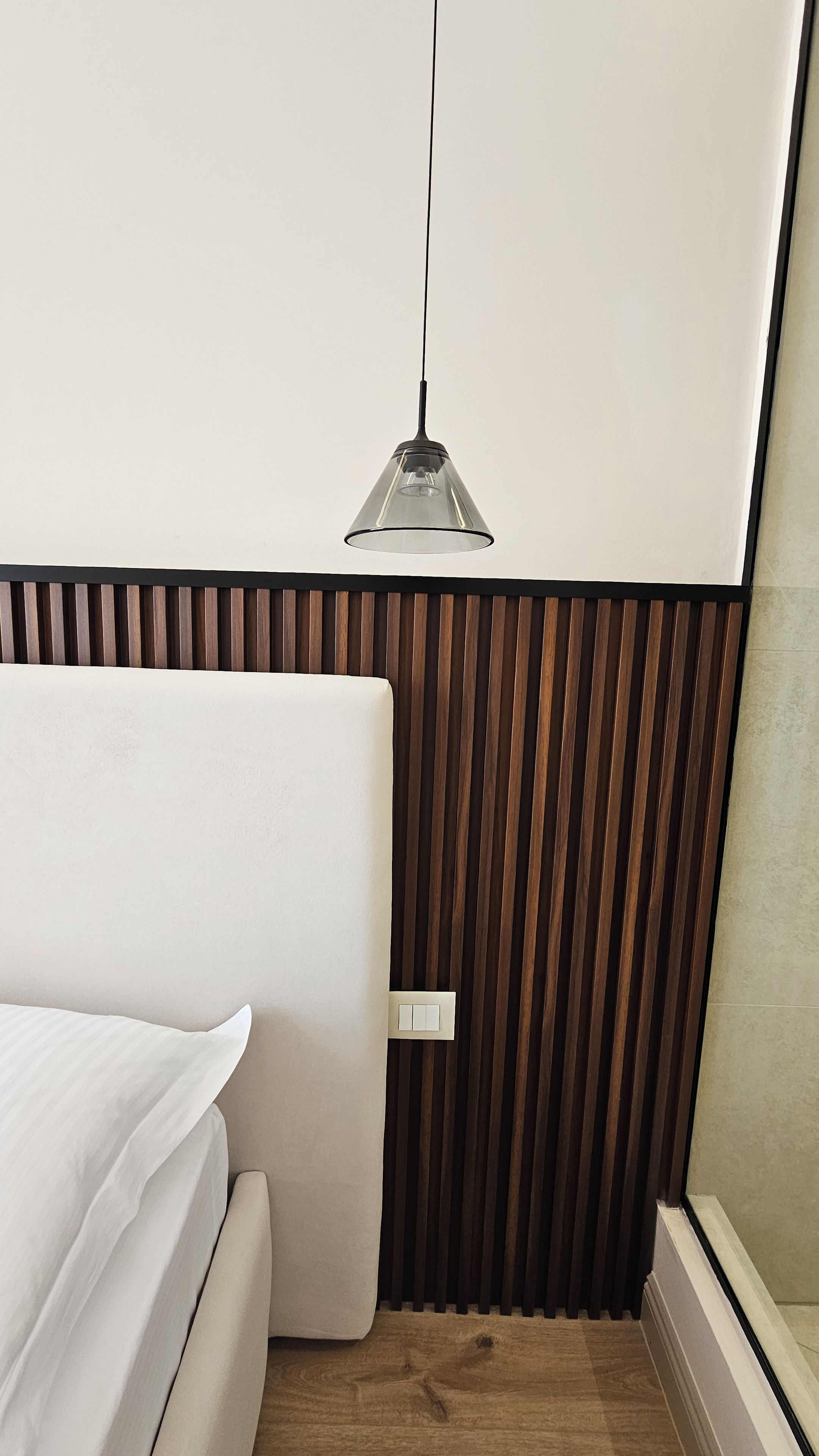 A room with a bed, a wooden slat wall, a hanging light fixture, and a switch on the wall.