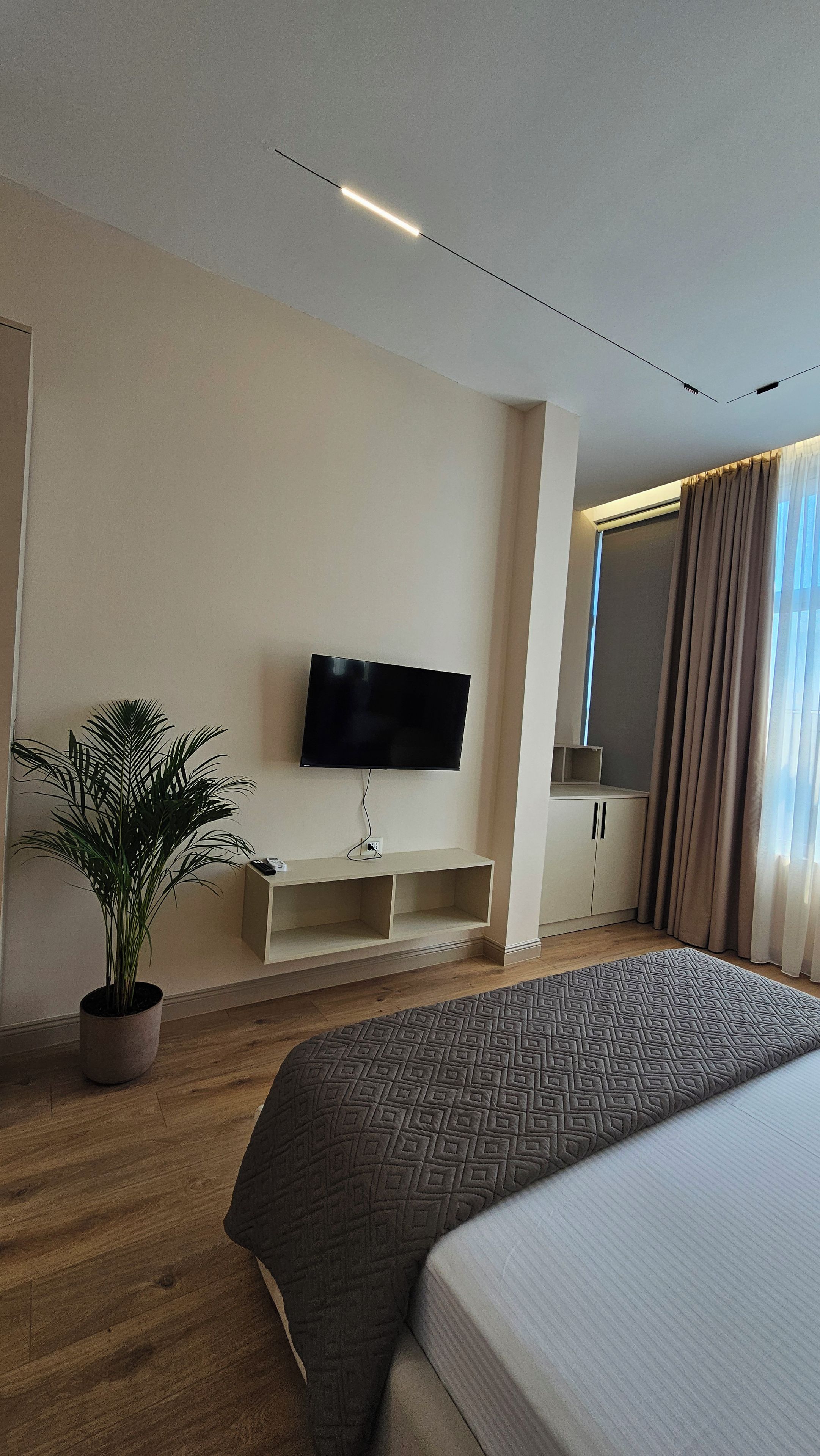 A modern hotel room with a flat-screen TV mounted on the wall, a potted plant, a wooden floor, and a bed with a patterned bedspread.