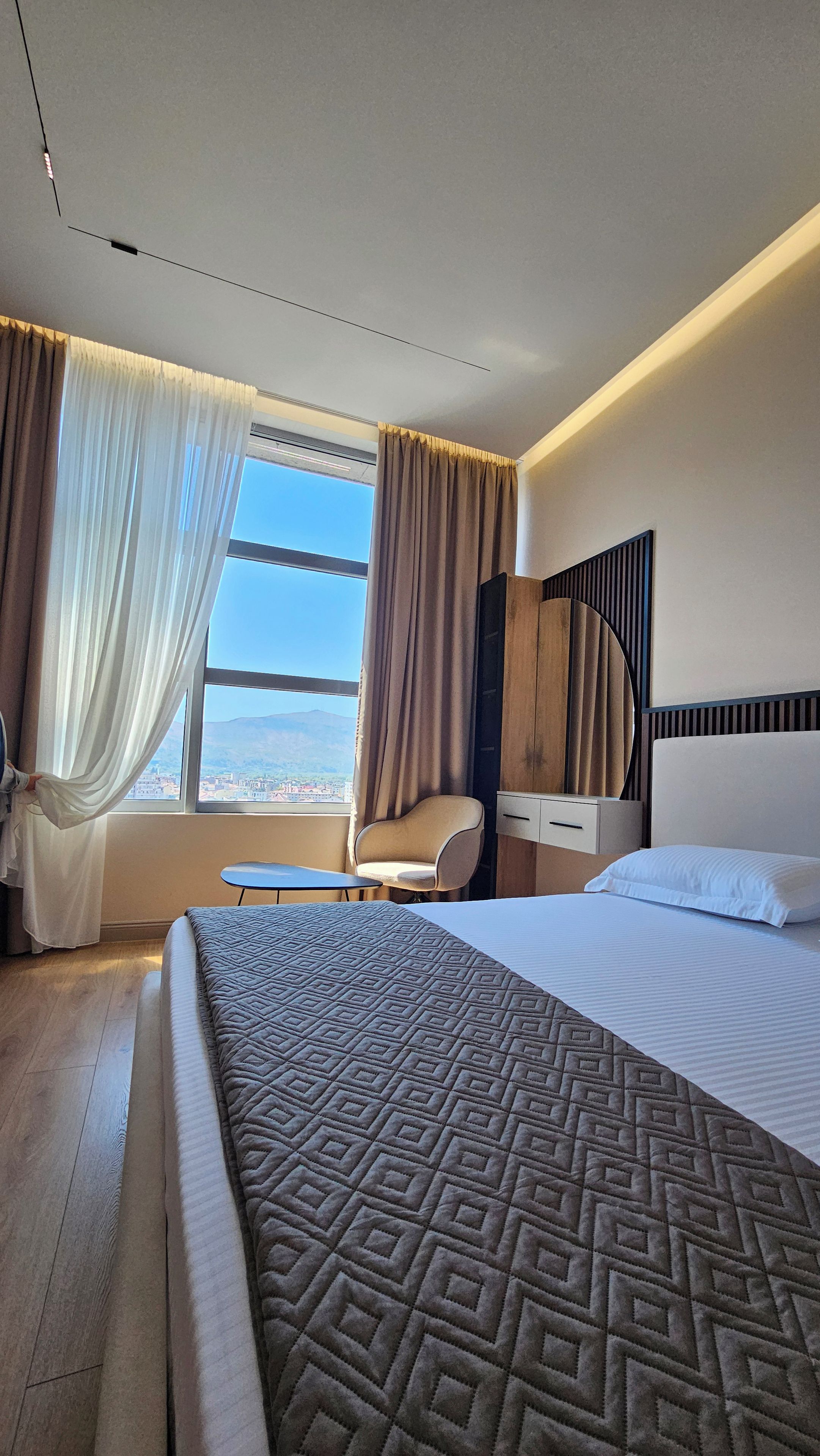 A hotel room with a large bed, a chair, a bedside table, and a view of the cityscape through the window.