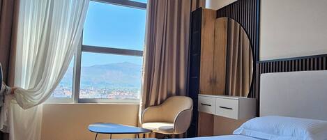 A hotel room with a large bed, a chair, a bedside table, and a view of the cityscape through the window.