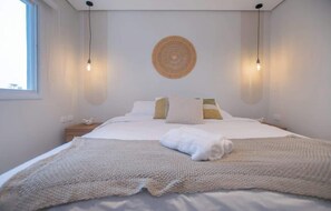 A neatly made bed with a white duvet and a knitted blanket, a bedside table with a small lamp, and a wall-mounted circular decorative item. - Corporate Stays First Living - 704 (Asunción)