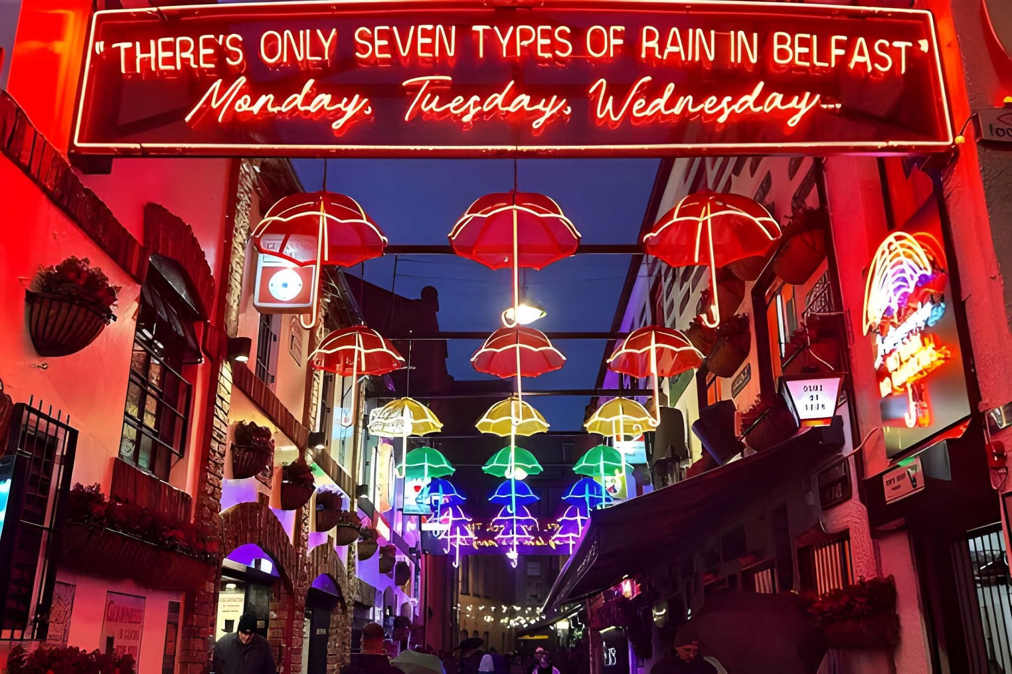Un letrero de neón con un mensaje humorístico sobre la lluvia en Belfast, y una calle con paraguas de colores colgando.