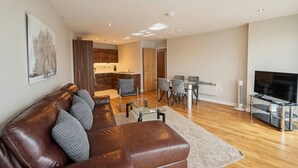 A modern living room with a leather sofa, a dining area with chairs, a kitchen in the background, and a flat-screen TV on the right. - Skyline & Sea (Belfast)