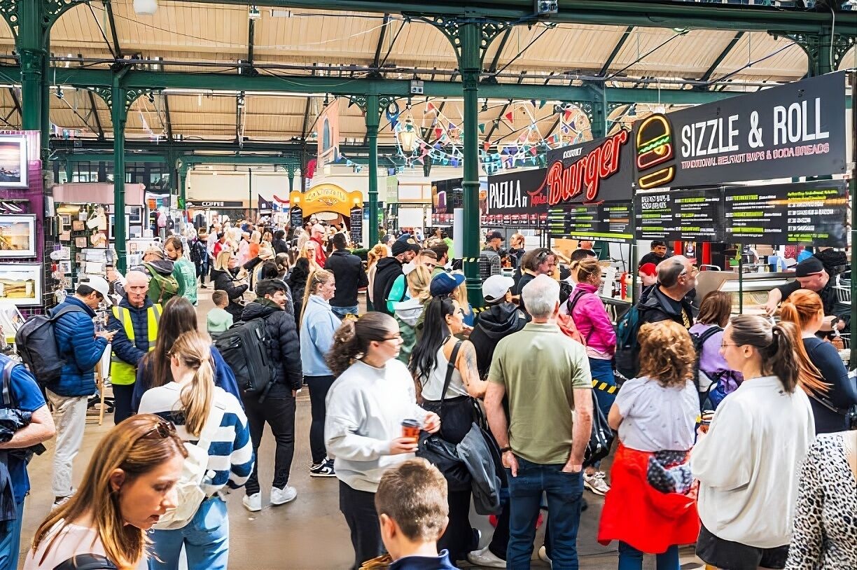 A crowded indoor market with various food stalls, including a burger stand and a sizzle & roll stand.