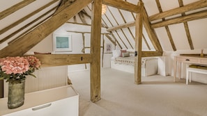 Quarto com vigas de madeira aparentes, cama com colcha florida, escrivaninha e um vaso com flores.
