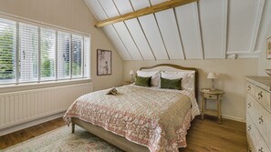 Quarto com teto inclinado, uma cama com colcha florida, mesa de cabeceira com abajur e cômoda.