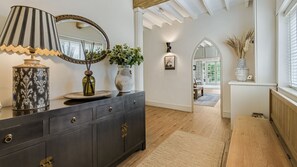 A hallway with a dark wooden dresser, a mirror, and a vase with plants. There is a door leading to another room with a window and a view of greenery outside. - The Old Bakery - sleeps 6 guests  in 3 bedrooms (Alderminster)