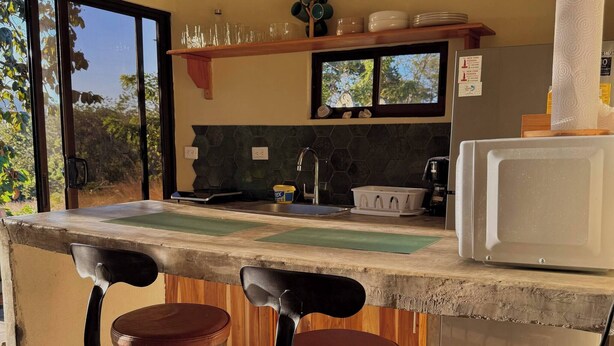 A kitchen with a bar area, two stools, a microwave, and a window. - Huella Cottage on Hilltop Views - Saltwater Pool (Cabo Velas)