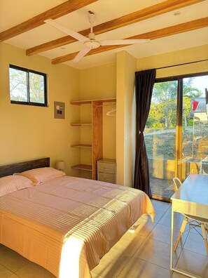 Ein Schlafzimmer mit Bett, Deckenventilator, Fenster, Kleiderschrank und Schreibtisch sowie Blick ins Freie.