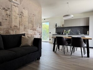 A modern living room with a black sofa, a dining area with a wooden table and chairs, and a kitchen with a black marble backsplash. - Apartment 8 – Palermo (Tremosine sul Garda)