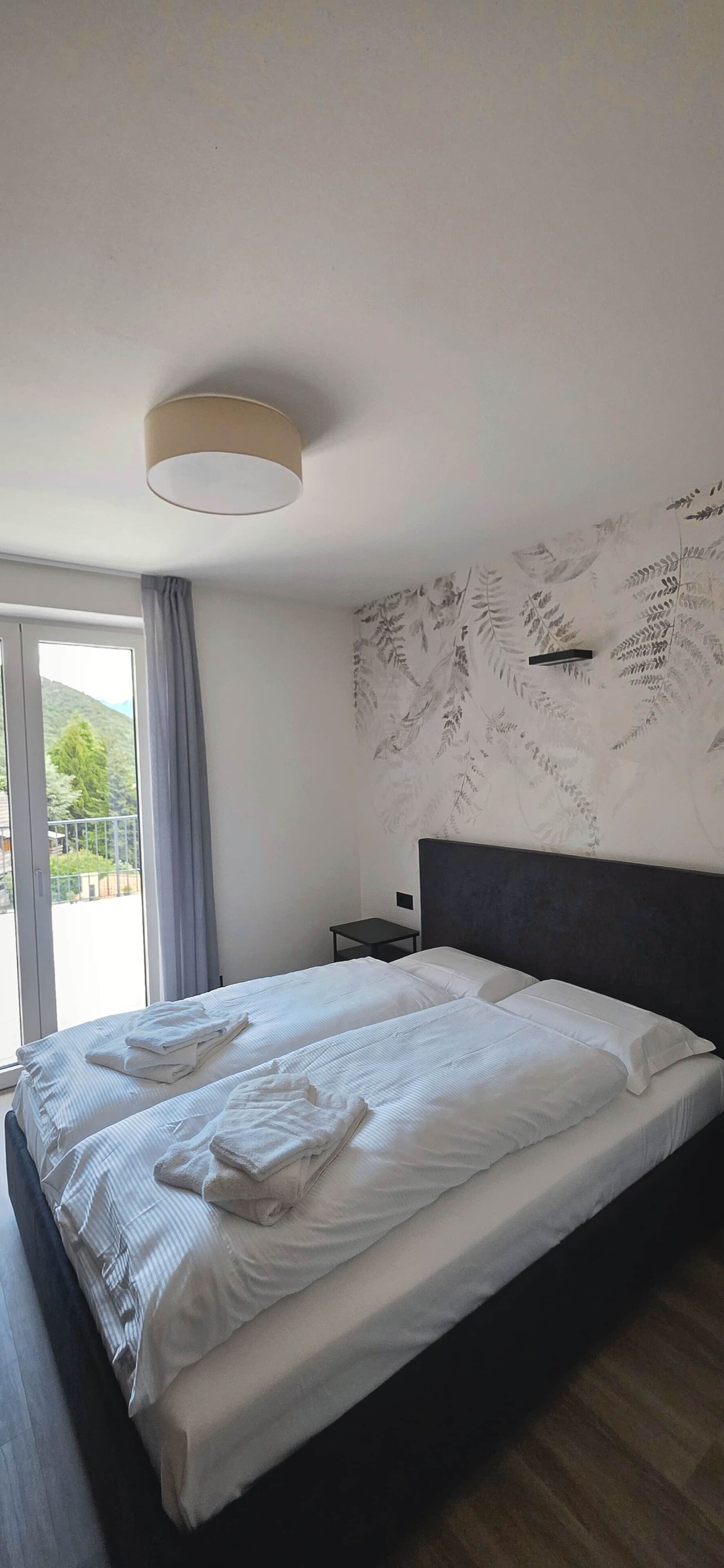Et hotelværelse med en seng, et natbord, en lampe, et vindue med gardiner og en væg med bladmotiver.