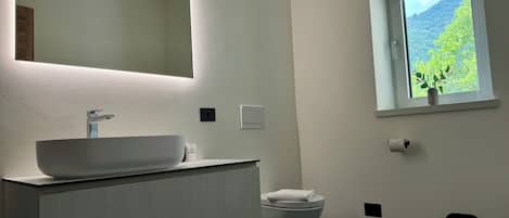 Une salle de bain moderne avec un grand miroir, un lavabo blanc, des toilettes et une fenêtre donnant sur un paysage verdoyant.