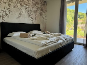 A bedroom with a large bed, a headboard, and a view of a green landscape through a sliding glass door. - Apartment 2 – Firenze (Tremosine sul Garda)