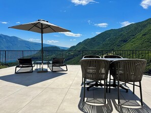 Eine Terrasse im Freien mit Tisch, Stühlen und einem großen Sonnenschirm, mit Blick auf eine schöne Berglandschaft.
