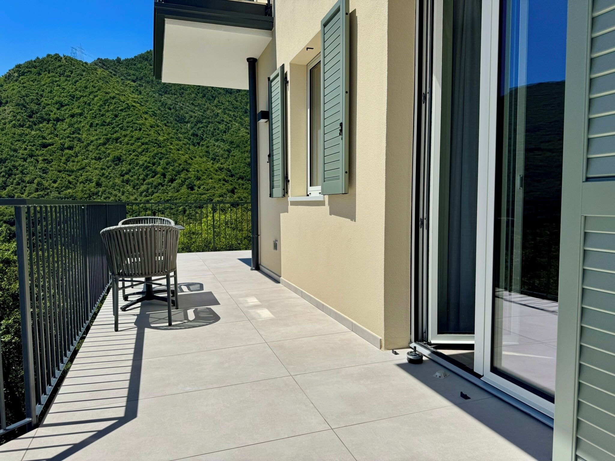 Ein Balkon mit einem Stuhl, ein Fenster mit Rollläden und Blick auf einen grünen Hügel.
