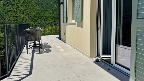 Ein Balkon mit einem Stuhl, ein Fenster mit Rollläden und Blick auf einen grünen Hügel.