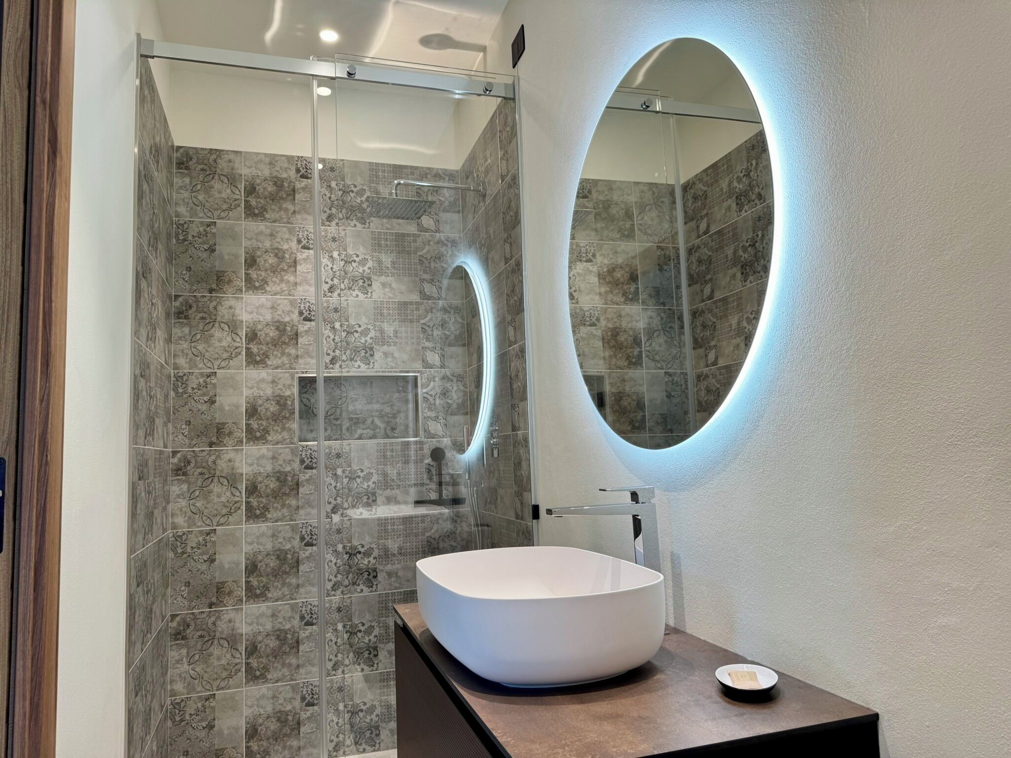 Ett modernt badrum med en duschavdelning med glasväggar, en vit tvättställ och en oval spegel som är monterad på väggen.