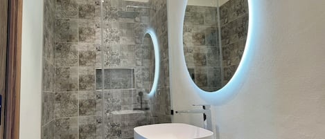 Un baño moderno con ducha envuelta en vidrio, un lavamanos blanco y un espejo ovalado fijado a la pared.