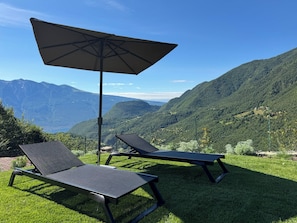 Zwei Liegestühle mit einem großen Sonnenschirm auf einer Rasenfläche mit Blick auf eine gebirgige Landschaft.