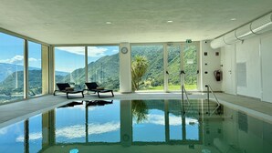 Ein Hallenbad mit großen Glaswänden, zwei Liegestühlen und Blick auf eine Berglandschaft.