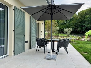 Un patio exterior con mesa y sillas, un paraguas grande y vistas a un jardín.