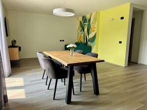 Ein Essbereich mit einem Holztisch, vier Stühlen, einer Vase mit Blumen sowie einer Wand mit Pflanzen- und Fruchtmotiv.