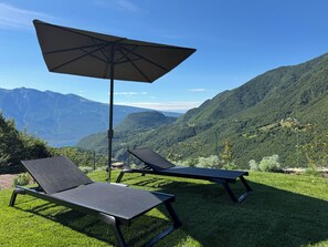 A pair of lounge chairs with a large umbrella on a grassy area, overlooking a mountainous landscape. - Apartment 7 – Limone (Tremosine sul Garda)