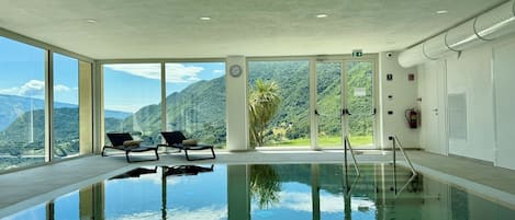An indoor swimming pool with clear glass walls, two lounge chairs, and a view of a mountainous landscape.