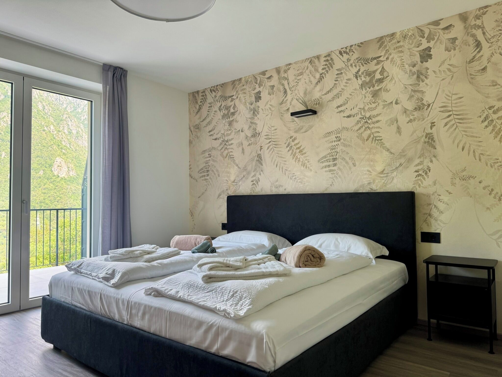 Une chambre avec un grand lit, une table de chevet et une vue sur la verdure par la fenêtre.