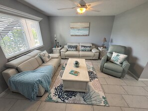 Un salón con un sofá beige, un sillón verde, una mesa de centro y un ventilador de techo.