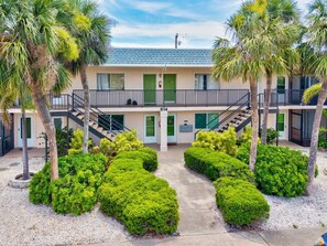 A two-story building with a balcony, a staircase, and a well-maintained garden with trimmed bushes and palm trees. - Cozy coastal 1-bedroom condo in lovely Venice Island (Venice)