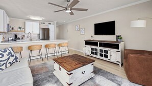 Una sala de estar moderna con sofá, una mesa de centro, televisor y una zona de cocina con taburetes de bar.