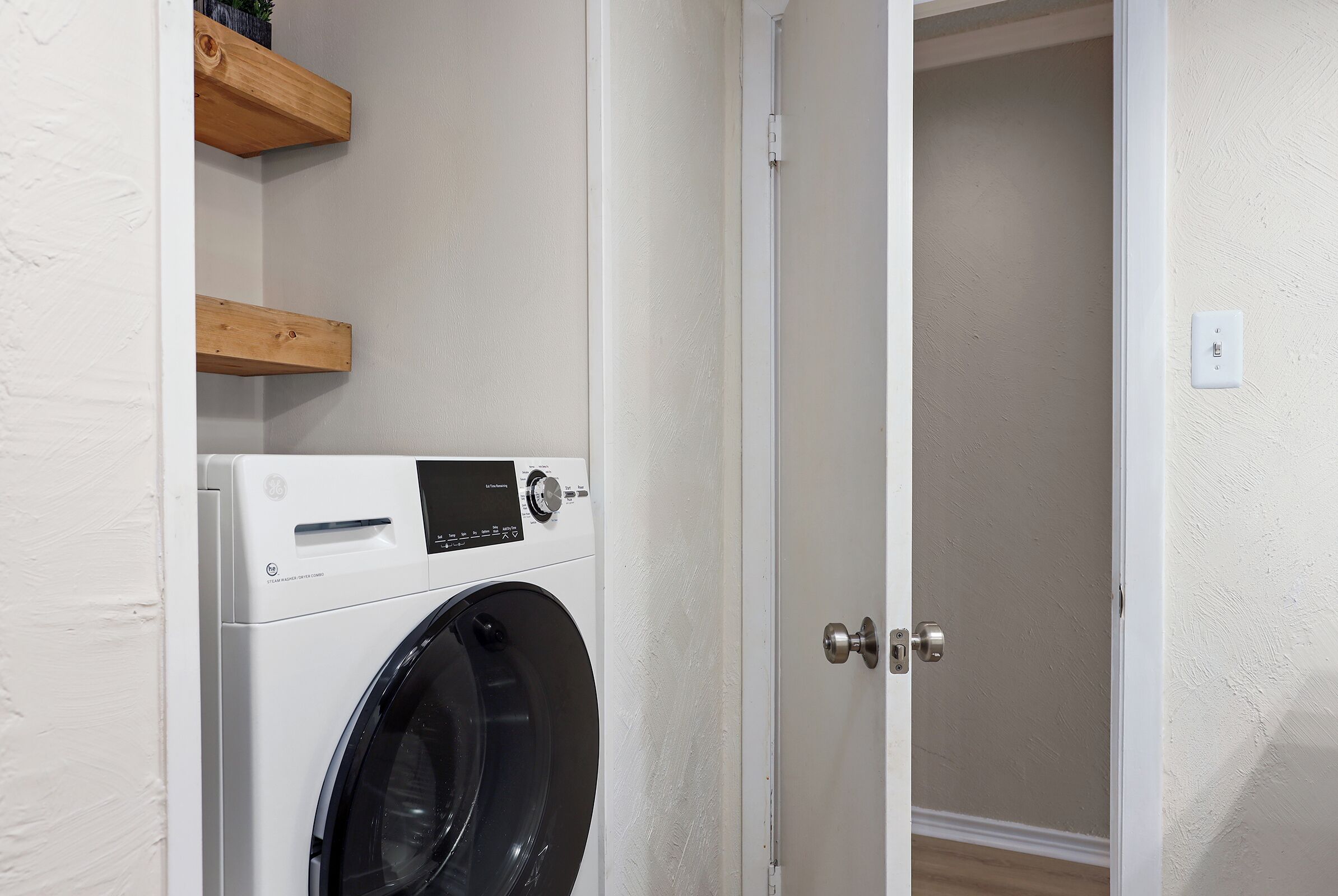 Een wasserette met een voorlader wasmachine, houten planken en een deur naar een andere ruimte.