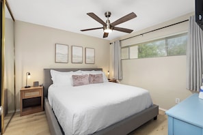 Une chambre à coucher avec un lit, un ventilateur de plafond, une table de chevet avec une lampe et une fenêtre avec des rideaux.