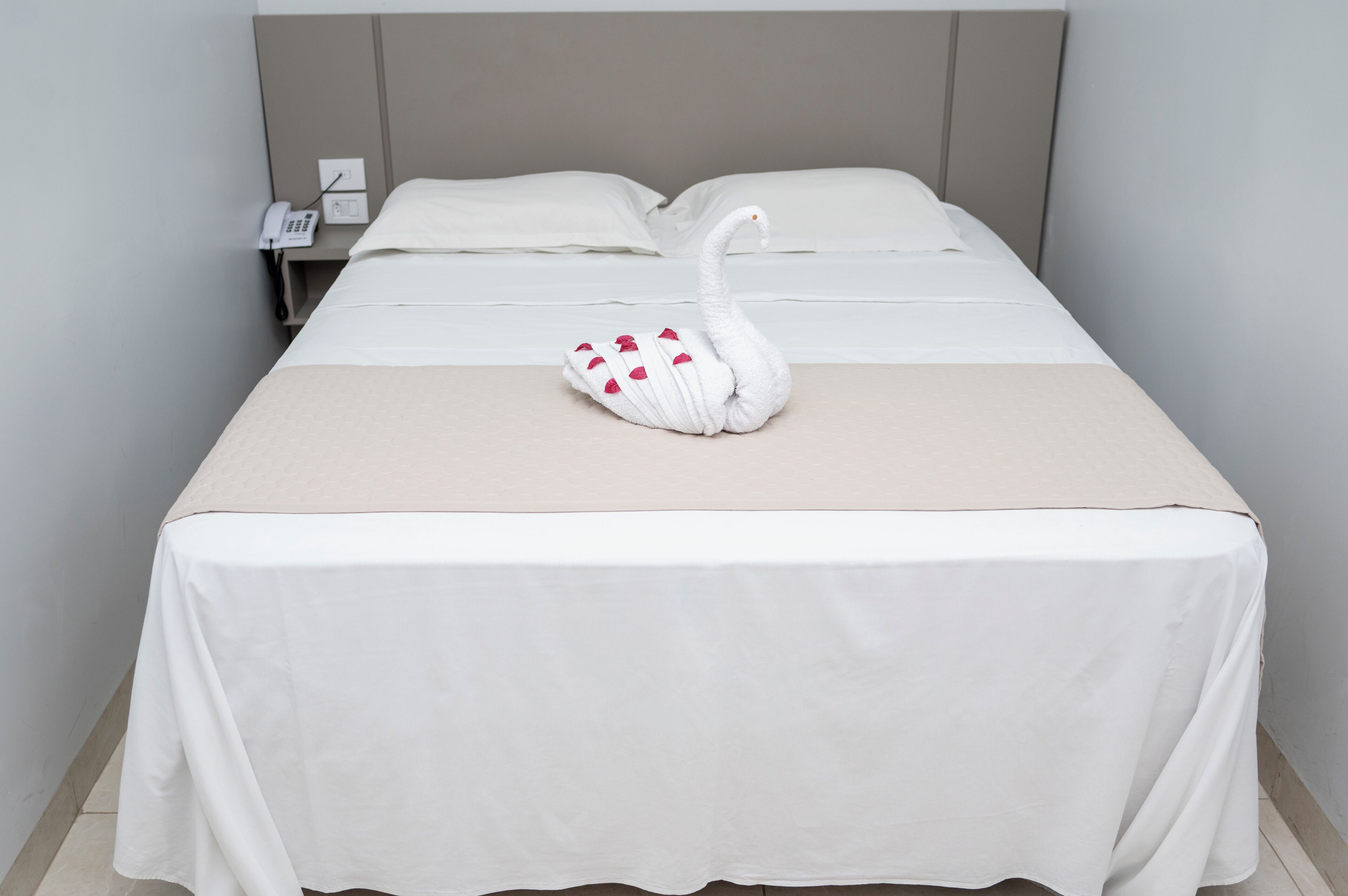 Ein ordentlich bezogenes Bett mit einem schwanenförmigen Kopfkissen, einem Nachttischtelefon und einer schlichten Wand.