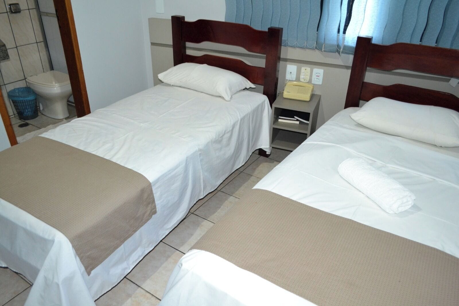 Une chambre d’hôtel avec deux lits simples, une table de chevet avec une lampe, une petite étagère et une fenêtre avec des rideaux.