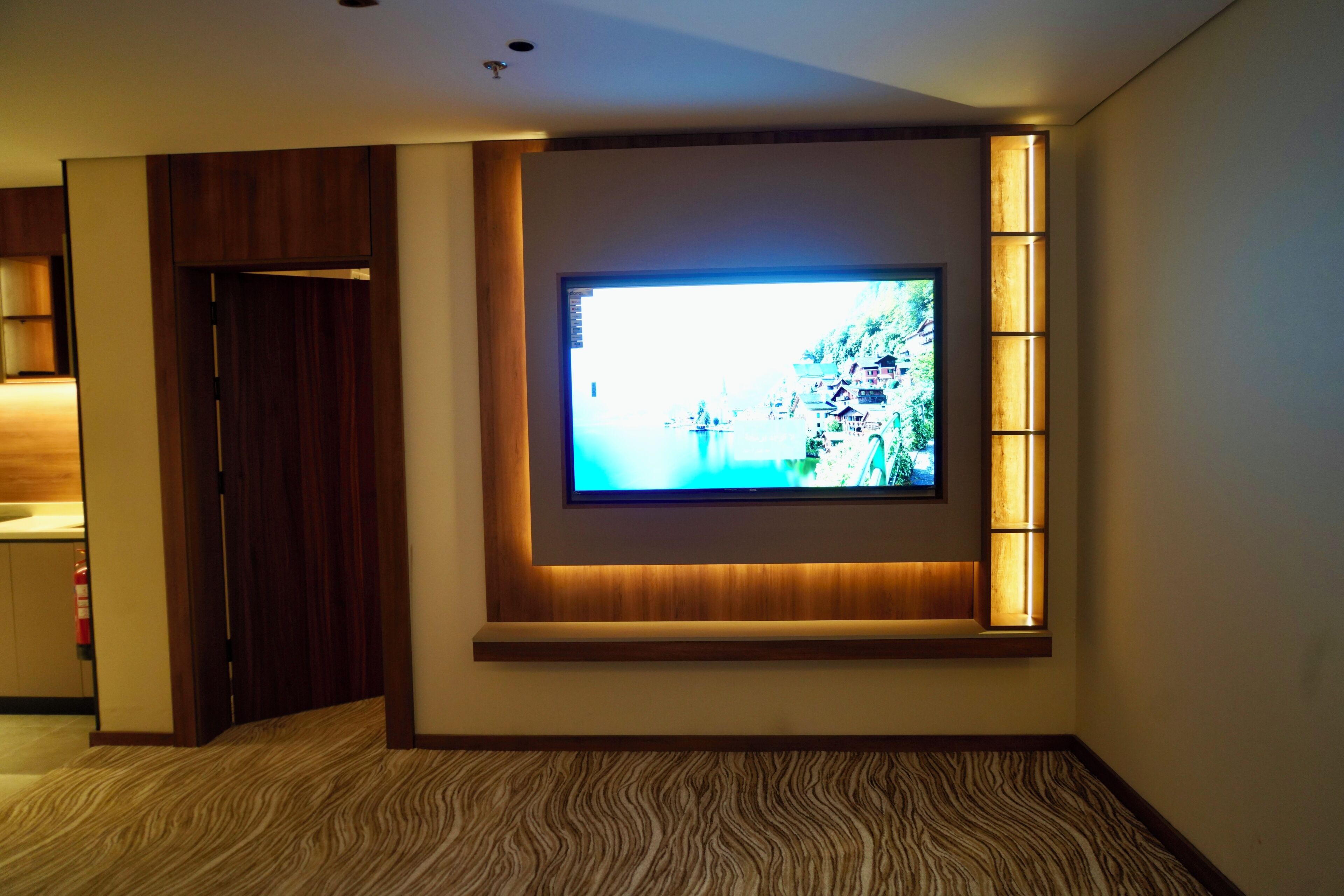 Quarto de hotel com uma televisão de tela plana grande fixada na parede, uma porta de madeira e um tapete estampado.