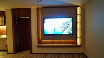 Quarto de hotel com uma televisão de tela plana grande fixada na parede, uma porta de madeira e um tapete estampado.