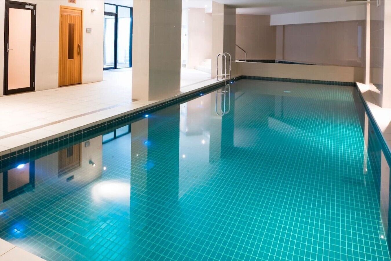 An indoor swimming pool with clear water, a ladder, and a tiled floor.