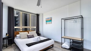 Ett modernt hotellrum med en stor säng, ett fönster med gardiner, en tavla, en svart metallhylla och en träbänk.