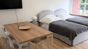 Une chambre moderne avec une table à manger en bois, deux lits, un téléviseur et une fenêtre donnant sur un mur de briques.