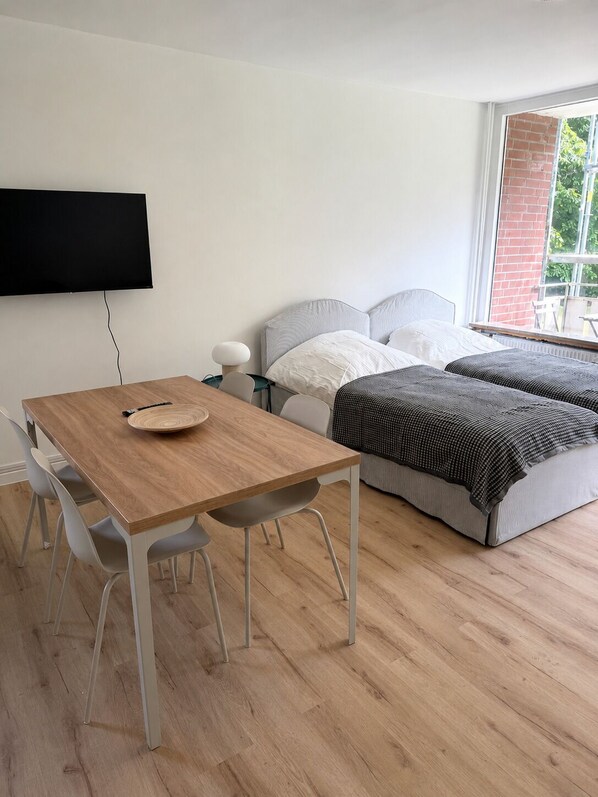 A modern bedroom with a wooden dining table, two beds, a TV, and a window with a brick wall view.