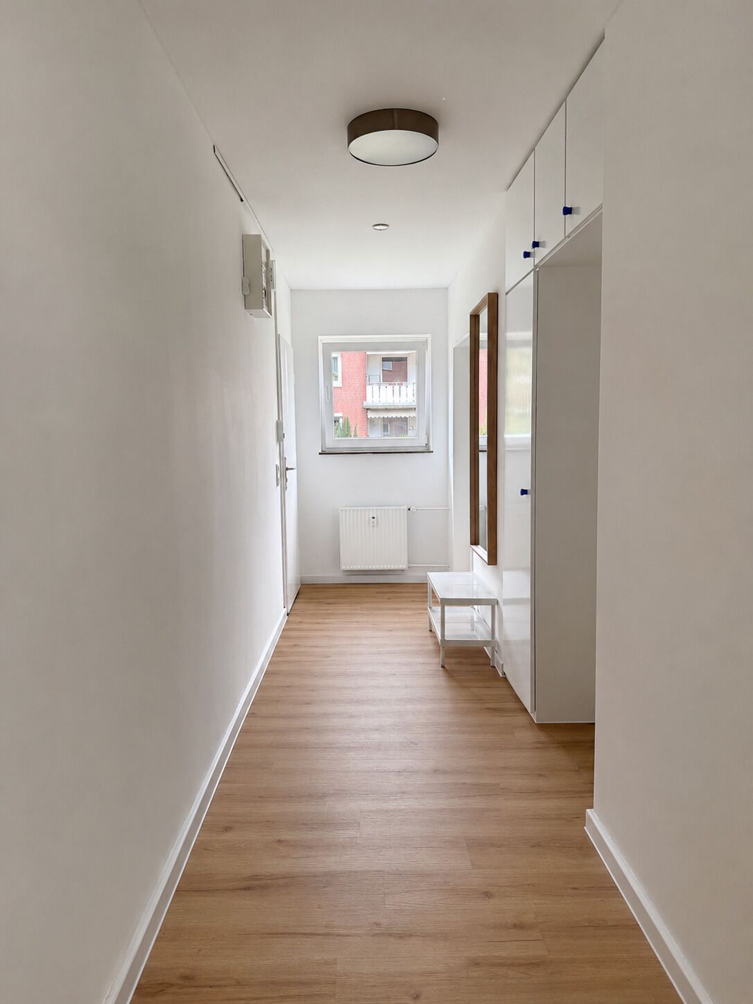 Ein schmales Dielenkorridor mit Holzboden, weißen Wänden, einem Fenster mit Blick auf ein Gebäude und einem weißen Heizkörper.