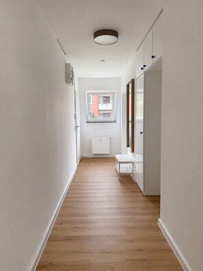 A narrow hallway with wooden flooring, white walls, a window with a view of a building, and a white radiator.