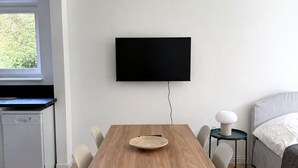 A modern room with a wooden dining table, two chairs, a flat-screen TV mounted on the wall, a small round table with a lamp, a bed with a white duvet, and a window with a view of greenery.