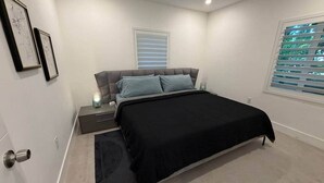 A bedroom with a bed, nightstand, lamp, and a window with blinds. - Home Private Pool • Next from the Design District (Miami)